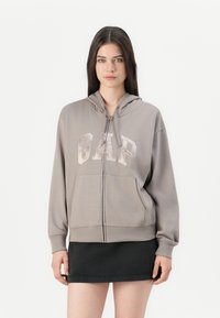 Kvinne i taupe zip-up hettegenser med "GAP"-logo og svart miniskjørt, stående mot en enkel hvit bakgrunn.