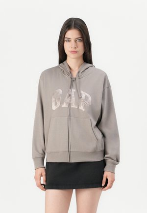 Žena u taupe zip-up majici s logom "GAP" i crnoj mini suknji, stoji protiv jednostavne bijele pozadine.