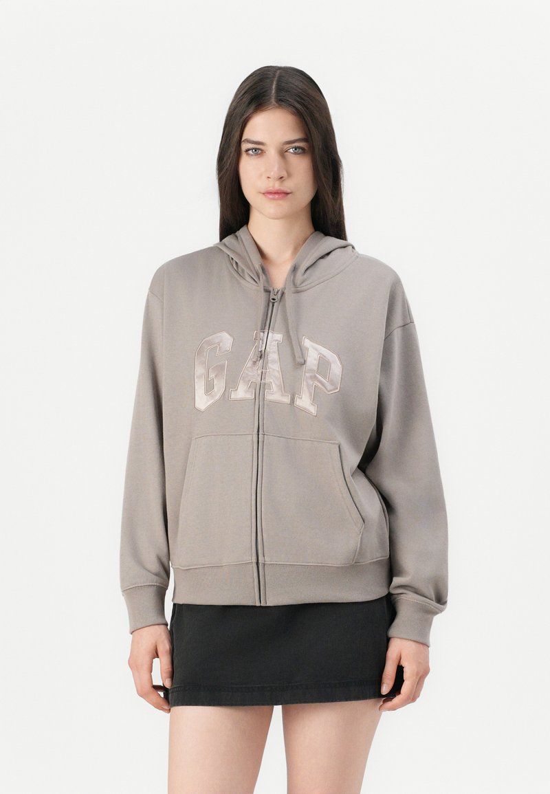 Kvinne i taupe zip-up hettegenser med "GAP"-logo og svart miniskjørt, stående mot en enkel hvit bakgrunn.