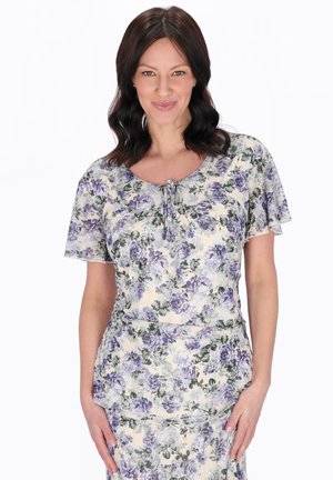 Kobieta o ciemnych włosach do ramion, ubrana w kremową sukienkę z fioletowym kwiatowym wzorem, stojąca i uśmiechająca się na białym tle.