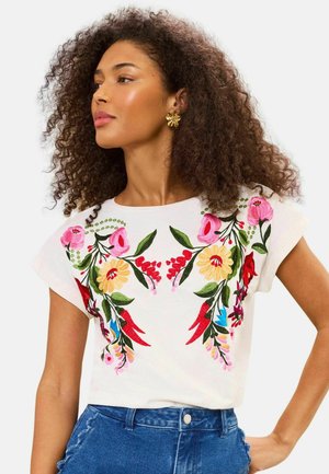 Kvinde med krøllet hår iført guldfarvede blomsterøreringe og en hvid top med farverig blomsterbroderi, kombineret med blå jeans.