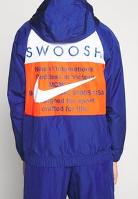 Barevná bunda Nike s modro-oranžovým designem, výrazným textem a logem, vyrobená z lehkého materiálu s kapucí.