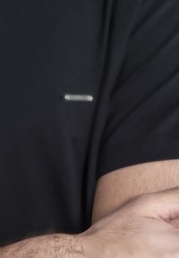 Čierne tričko s krátkym rukávom vyrobené z mäkkej tkaniny, s malým strieborným logom na oblasti hrude a voľným strihom okolo rúk.