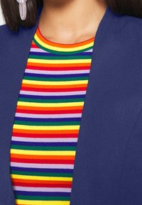 Haut rayé multicolore en rouge, jaune, vert, bleu et violet, associé à une veste unie bleu marine. Tissu à texture lisse, coupe ajustée.