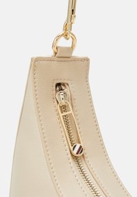 Szczegół beżowej torby skórzanej z złotym zamkiem błyskawicznym, teksturowaną powierzchnią i okrągłym uchwytem zamka z paskowanymi akcentami.