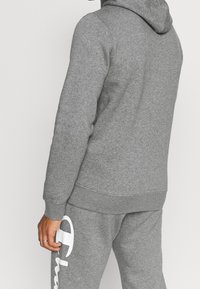 Sudadera gris con capucha y puños acanalados, hecha de un material suave. Presenta un gran logo blanco en la parte inferior. Combinada con pantalones de chándal a juego.