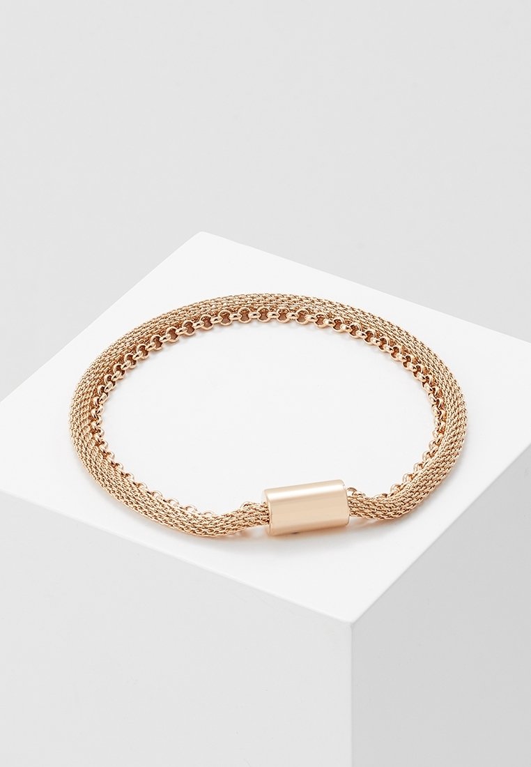 Bracelet chaîne en maille dorée avec fermoir magnétique cylindrique sur une surface blanche inclinée sur un fond gris clair.