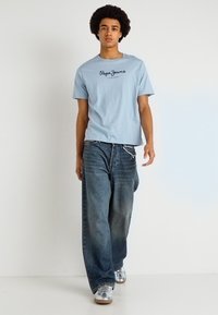 Ανοιχτό μπλε βαμβακερό μπλουζάκι με κοντά μανίκια και μαύρο λογότυπο κειμένου; συνδυασμένο με φαρδιά, ξεθωριασμένα μπλε τζιν παντελόνια και ασημί αθλητικά παπούτσια.