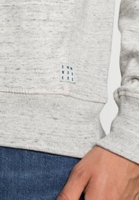 Sudadera gris claro con una tela texturizada, puños y dobladillo canalé. Presenta una etiqueta azul con "DNM WORKR" en el lateral inferior.