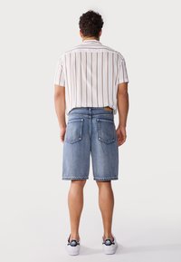 Wit gestreept overhemd met korte mouwen, blauwe denim short en witte sneakers. Opvallende kenmerken zijn de button-down kraag en twee achterzakken.