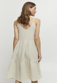 Cremfarbendes, ärmelloses Kleid mit V-Ausschnitt und gestuftem Saum. Hergestellt aus strukturiertem Stoff und in entspannter Silhouette.