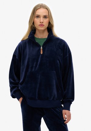 Mørkeblå, fløjlsagtig pullover med høj krave, halvlynlås og frontlomme. Har et subtilt logo på brystet og en afslappet pasform.