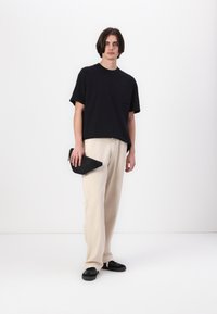 Zwarte t-shirt met korte mouwen en een borstzak, gecombineerd met crème kleurige broek met wijde pijpen en zwarte instappers. Houdt een zwarte clutch vast.