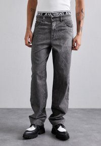 Jeans din denim gri cu un finisaj texturat și cus�ături vizibile, având un braguard negru Versace. Asortați cu pantofi negri și albi.