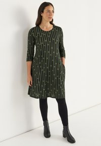 Vihreä mekko mustalla viivakuviolla, 3/4-hihat, sivutaskut ja pyöreä pääntie, yhdistetty mustiin leggings-vaatteisiin ja nilkkabootseihin.