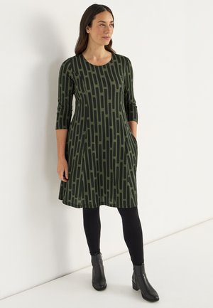 Groene jurk met zwart lineair patroon, 3/4 mouwen, zijvakken en een ronde halslijn, gecombineerd met zwarte leggings en enkellaarsjes.