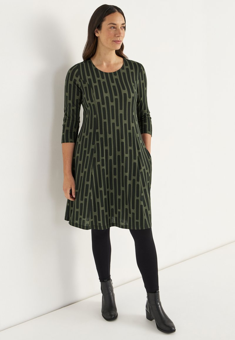Vihreä mekko mustalla viivakuviolla, 3/4-hihat, sivutaskut ja pyöreä pääntie, yhdistetty mustiin leggings-vaatteisiin ja nilkkabootseihin.