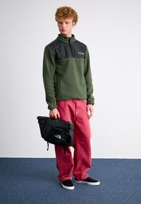 Fleece'owy pulower w ciemnozielonym kolorze z czarnym panelem na klatce piersiowej, w zestawieniu z luźnymi czerwonymi spodniami i czarną torbą na ramię. Czarne buty wsuwane.