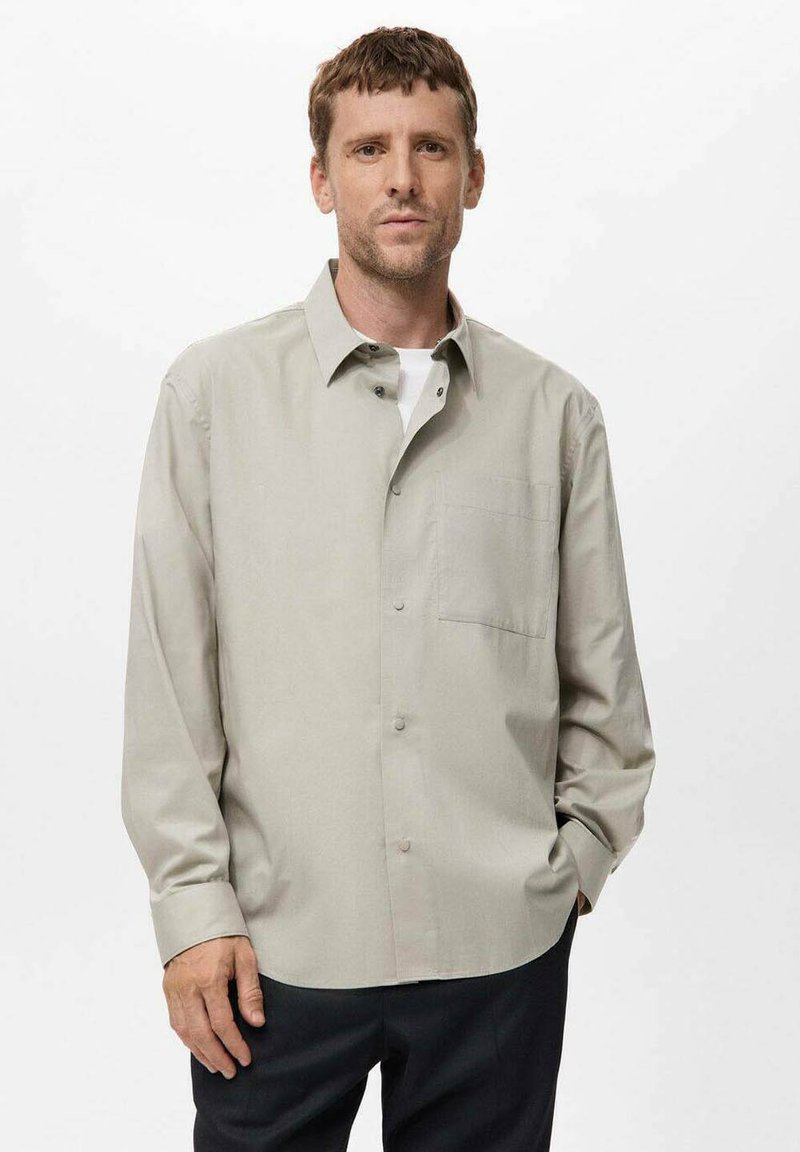 Mann mit kurzen braunen Haaren, der ein beigefarbenes Hemd mit Knöpfen über einem weißen T-Shirt trägt und vor einem schlichten weißen Hintergrund steht.