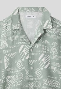 Camicia a maniche corte di colore verde chiaro con motivi geometrici bianchi e una texture morbida. Presenta un colletto classico e bottoni sulla parte anteriore.