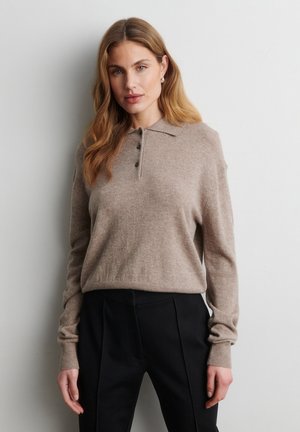 Vrouw met lang golvend haar, die een beige trui met lange mouwen en knopen en een zwarte broek draagt, staand tegen een eenvoudige lichte muur.