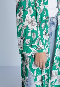 Lang, flydende kimono i grøn stof med hvide blomsterprint. Har løse ærmer og et spaltet design, parret med blå denimjeans.