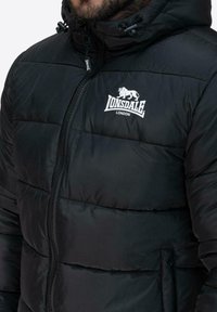 Czarna kurtka puchowa z zamkiem błyskawicznym, regulowanym kapturem i logo Lonsdale. Posiada pikowaną fakturę i elastyczne mankiety dla dodatkowego ciepła.