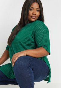 Grön texturerad topp med horisontella ränder, sidofickor och korta ärmar, ihop med mörkblå denimjeans.