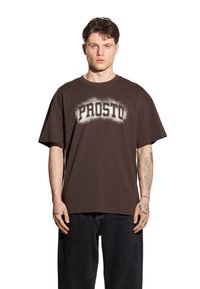 Brązowy t-shirt wykonany z bawełny z napisem "PROSTO" w białych, postarzanych literach na piersi; luźny krój i krótkie rękawy.