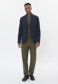 Blazer blu navy sopra un maglione grigio a maglia, abbinato a chinos oliva e scarpe marroni. Il look presenta linee pulite e vestibilità classiche.