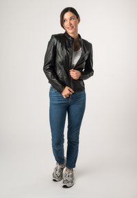 Schwarze Lederjacke mit schmalem Schnitt, hohem Kragen und Reißverschluss. Kombiniert mit blauen Jeans und grauen Sneakers vor einem neutralen Hintergrund.