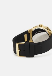 Musta silikoni kello hihna, jossa on kultainen metallipidike. Pyöreä kellotaulu kiiltävällä pinnalla, teksturoidulla takapuolella ja minimalistisella muotoilulla.