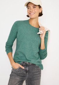 Maglione verde in lana con scollo rotondo e maniche lunghe, abbinato a jeans grigi. La modella tiene un oggetto beige su una spalla.