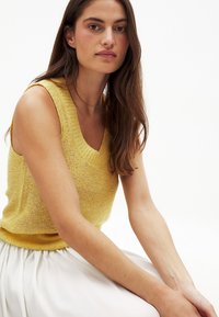 Haut sans manches en tricot jaune avec un décolleté en V, présentant un motif texturé. Le mannequin a les cheveux longs et est assis contre un fond blanc uni.