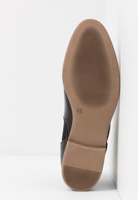 Semelle de chaussure en cuir noir avec semelle en caoutchouc marron clair, taille 37 indiquée, positionnée à la verticale contre un mur blanc d'angle.