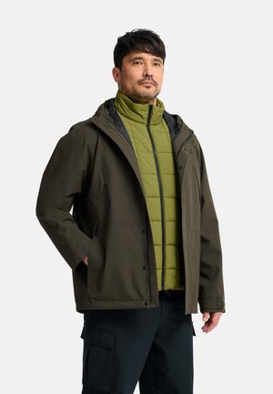Man die gelaagde outdoorjassen draagt, een olijfgroene pufferjas onder een donkergroene waterdichte jas, staand met handen in de zakken.