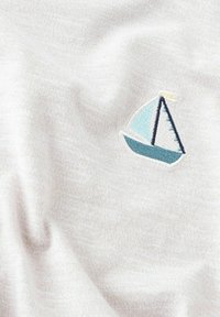 Tela blanca con una textura suave que presenta un pequeño velero bordado en azul claro y azul oscuro, con un acento amarillo en la vela.
