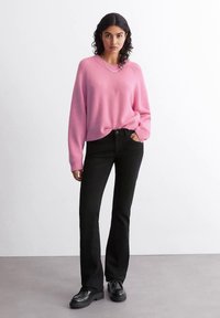 Γυναίκα με μαύρα σγουρά μαλλιά, φορώντας ροζ πουλόβερ, μαύρα τζιν, μαύρα παπούτσια, στέκεται με το ένα χέρι στην τσέπη της, μπροστά σε απλό φόντο.