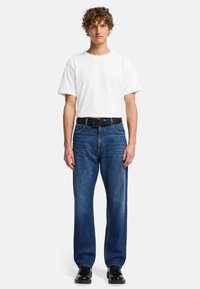 Witte katoenen t-shirt, blauwe denim recht-valle spijkerbroek, zwarte riem met zilveren gesp, en zwarte schoenen. Model staat tegen een effen achtergrond.
