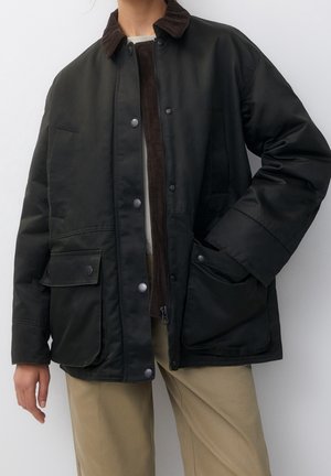 Persona con chaqueta negra abotonada con grandes bolsillos delanteros sobre un suéter marrón con cremallera y pantalones beige sobre un fondo blanco.