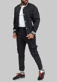 Veste bombardier noire avec col rayé blanc, boutons blancs, associée à un pantalon cargo noir avec poches et des baskets noires avec des accents orange.