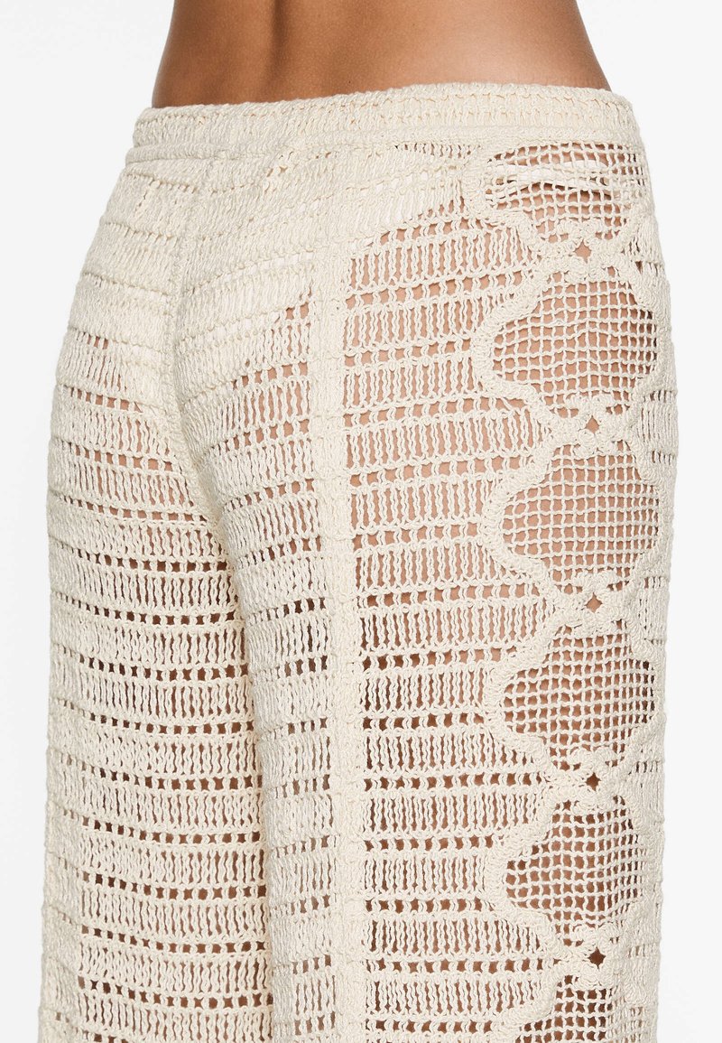 Vue de côté d'une personne portant un pantalon en crochet beige avec un motif floral en forme de volute et des détails en maille ouverte sur la peau.