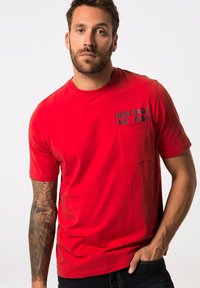 Camiseta de algodón roja con mangas cortas, que presenta un bolsillo y "HERITAGE IND. 1880" impreso en negro. El modelo tiene el brazo tatuado.