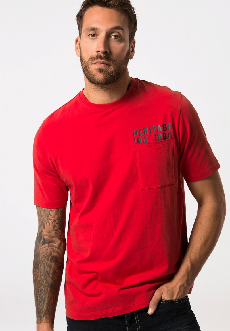 Camiseta de algodón roja con mangas cortas, que presenta un bolsillo y "HERITAGE IND. 1880" impreso en negro. El modelo tiene el brazo tatuado.