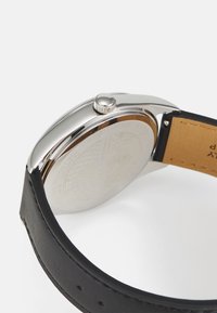 Montre en acier inoxydable argenté avec une finition polie, bracelet en cuir noir et un fond de boîtier texturé avec des détails gravés.