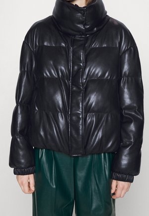 Veste en cuir noire matelassée avec col haut et boutons-pression portée sur un pantalon en cuir vert foncé, montrée de la taille au cou.