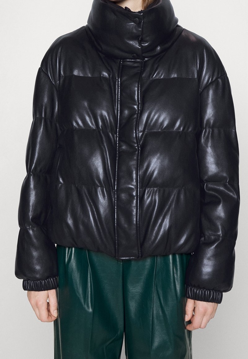 Veste en cuir noire matelassée avec col haut et boutons-pression portée sur un pantalon en cuir vert foncé, montrée de la taille au cou.