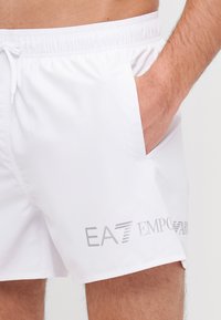 Șorturi de baie albe dintr-un material fin, având talie elastică, buzunar lateral și un logo gri EA7 Emporio Armani pe lateral.