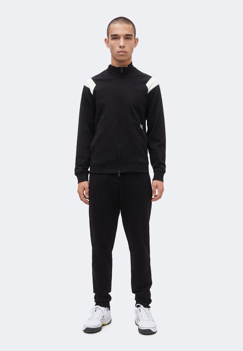 Νέος άνδρας όρθιος, φορώντας μαύρο μπουφάν με φερμουάρ και λευκά σημεία στους ώμους, μαύρο παντελόνι που ταιριάζει και λευκά αθλητικά παπούτσια σε ανοιχτό φόντο.