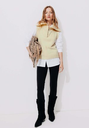Gilet en tricot zippé de couleur jaune pâle, superposé à une chemise blanche, associé à un legging noir et à de grandes bottes de cowboy noires. Accessoirisé avec un sac à motif léopard.
